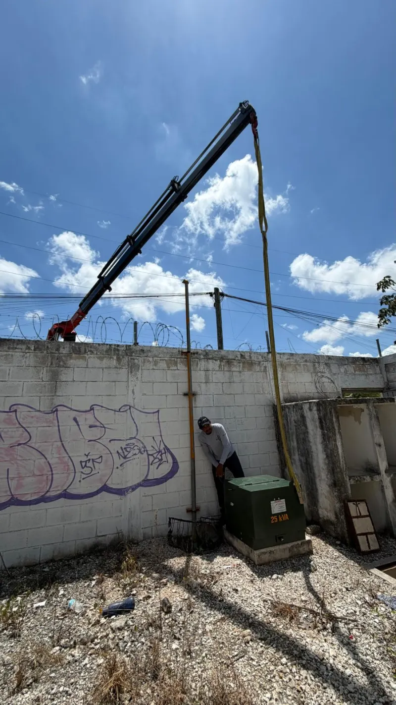 Transformador verde sobre una base de madera, suspendido con dos eslingas frente a la grúa.