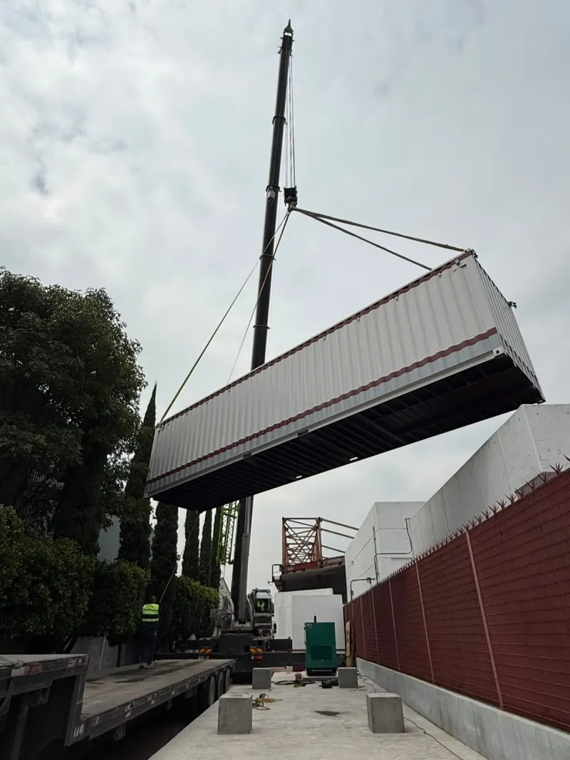 Contenedor blanco suspendido de una grúa telescópica sobre un pasillo entre árboles y una barda roja.