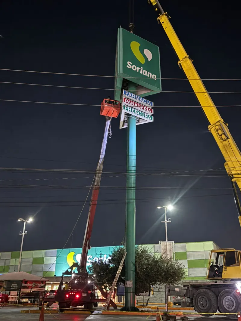 Tótem verde con anuncio superior y letreros inferiores en un estacionamiento, con vehículos y líneas aéreas.