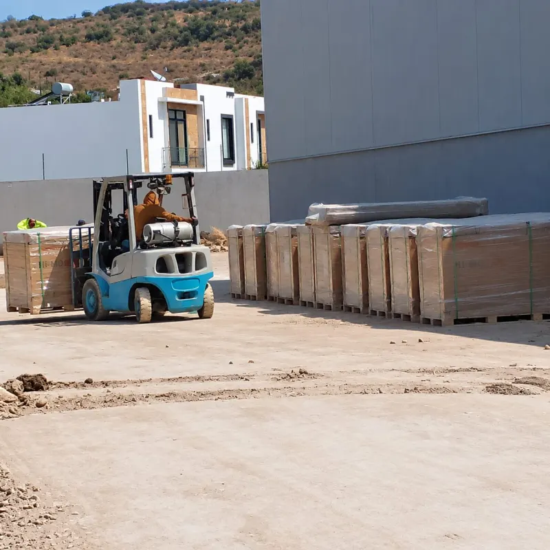 Montacargas azul transporta una caja embalada junto a otras cajas sobre tarimas frente a la nave.