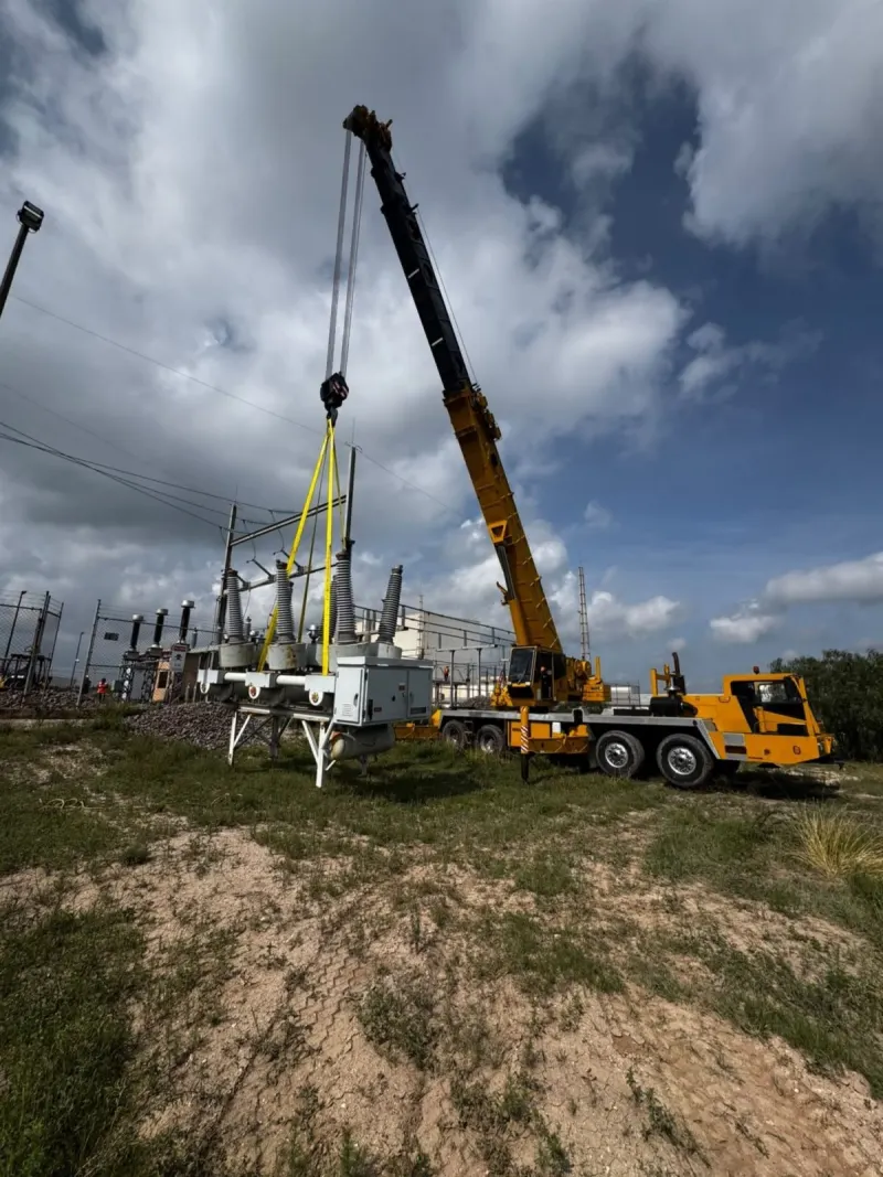 Equipo eléctrico gris con aisladores, suspendido por eslingas amarillas dentro de la subestación.