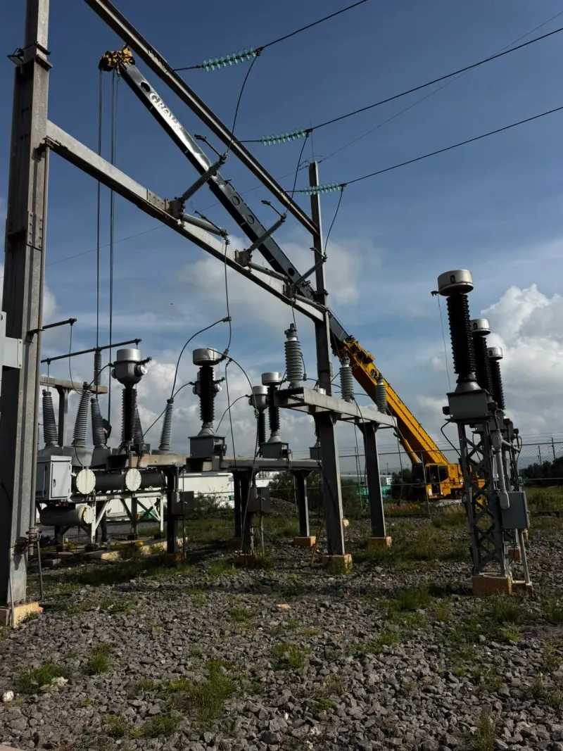 Grúa telescópica amarilla eleva un equipo eléctrico gris con eslingas dentro de la planta.